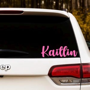 May include: A white Jeep SUV with a pink vinyl decal that reads "Kaitlin" in a cursive font on the rear window. The taillight is red, and the background shows fall foliage.
