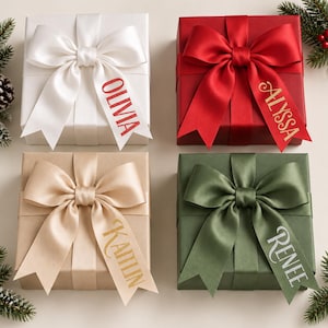 May include: Four square gift boxes wrapped in solid white, red, tan, and olive green paper. Each box features a large satin ribbon bow with a personalized name printed on one tail: Olivia in red, Alyssa in gold, Kaitlin in gold, and Renee in white.