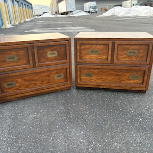 May include: Two wooden chests of drawers in a warm brown finish. Each chest has two small drawers above a larger drawer, all with brass-toned handles. The chests are rectangular and sit on a low base.