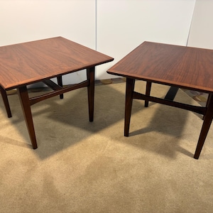 Puede incluir: Dos mesas auxiliares cuadradas de estilo moderno de mediados de siglo. Las mesas tienen un acabado de madera marrón oscuro con patas anguladas y una viga de soporte inferior. Las mesas miden aproximadamente 61 cm de ancho y 61 cm de largo.
