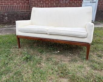 May include: A cream-colored two-seater sofa with a wooden frame and legs. The sofa has a high back and armrests. The cushions are a lighter shade of cream. The sofa is set on a grassy lawn.