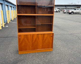 May include: A large, mid-century modern wooden cabinet with a hutch. The upper section has glass sliding doors and shelves. The lower section features wooden sliding doors. The cabinet is made of light brown wood.