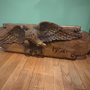 May include: A rectangular wooden wall plaque featuring a carved eagle with outstretched wings in the centre. The wood has a dark brown stained finish with a textured surface. The number 1776 is carved into the lower right side of the plaque.