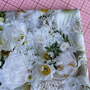 Puede incluir: Primer plano de una tela con un estampado floral. El diseño presenta una variedad de flores blancas, incluyendo peonías y margaritas, con centros amarillos y follaje verde. La tela está doblada, mostrando el estampado detallado.