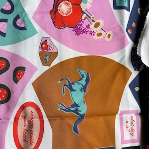 Puede incluir: Una tela blanca con un patrón colorido. El patrón incluye rodajas de sandía, un caballo azul, una flor roja y texto que dice "grita dentro de tu corazón" y "¿qué día es?"