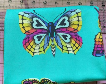 May include: A folded turquoise fabric featuring a large, colorful butterfly print. The butterfly has yellow, orange, purple, and black details. The fabric is on a pink cutting mat with measurement markings.