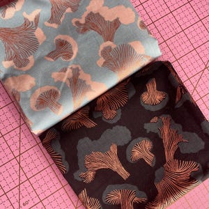 May include: Two folded fabric pieces with mushroom patterns. One fabric is light blue with pink and copper-colored mushrooms. The other is dark brown with gray and copper-colored mushrooms. The fabrics are on a pink cutting mat.