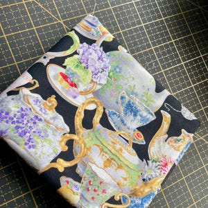 May include: Folded black fabric with a repeating pattern of ornate teacups, teapots, and floral arrangements in shades of white, blue, green, and purple. Gold accents are visible on the teapots. The fabric is folded on a black cutting mat.