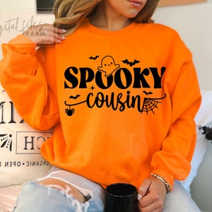 Pode incluir: Camisola laranja com texto preto que diz "Spooky Cousin" e um gráfico de fantasma.