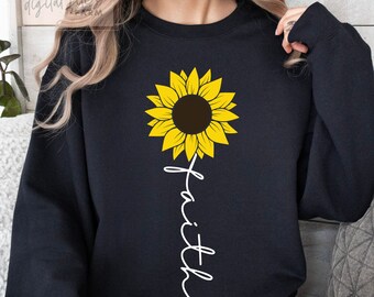 Può includere: Felpa nera con un'immagine di girasole giallo e la parola "fede" scritta verticalmente sotto.