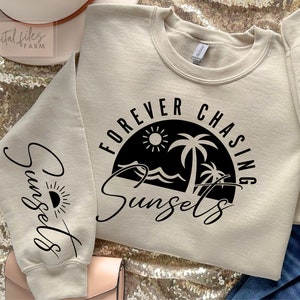 Könnte beinhalten: Ein beiger Pullover mit schwarzem Text, der "Forever Chasing Sunsets" in einem Kreis mit einer Palme und einem Sonnen-Motiv zeigt. Der Text "Sunsets" ist auch auf dem Ärmel des Pullovers geschrieben.