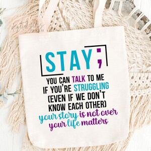 K&ouml;nnte beinhalten: Eine wei&szlig;e Einkaufstasche mit dem Text "STAY; YOU CAN TALK TO ME IF YOU'RE STRUGGLING (EVEN IF WE DON'T KNOW EACH OTHER) your story is not over your life matters" in Schwarz, Blau und Lila gedruckt.