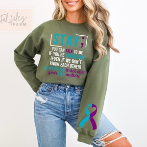 K&ouml;nnte beinhalten: Gr&uuml;ner Pullover mit dem Text "STAY; YOU CAN TALK TO ME IF YOU'RE STRUGGLING (EVEN IF WE DON'T KNOW EACH OTHER) YOUR STORY IS NOT OVER YOUR LIFE MATTERS" in T&uuml;rkis und Lila. Der Pullover hat ein t&uuml;rkisfarbenes und lilafarbenes Band am &Auml;rmel.