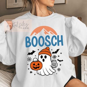 Op de afbeelding: Witte sweatshirt met een Halloween-thema. Het ontwerp toont het woord "BOOSCH" in blauw, een cartoongeest met een oranje muts, die een pompoen en een drankje vasthoudt. Zwarte vleermuizen, spinnenwebben en sterren maken het ontwerp compleet.