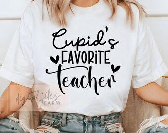 以下が含まれることがあります： 黒い文字で「Cupid's Favorite Teacher」と書かれた白いTシャツ。テキストの両側にハートマークがあります。