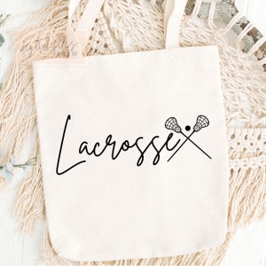 Może przedstawiać: Biała torba z pł&oacute;tna z czarnym tekstem, kt&oacute;ry m&oacute;wi "Lacrosse" i parą kij&oacute;w do lacrosse z piłką w środku.