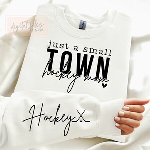 Może przedstawiać: Biała bluza z czarnym napisem "Just a small town hockey mom" z małym symbolem serca. Rękaw bluzy ma napis "Hockey X" w kolorze czarnym.