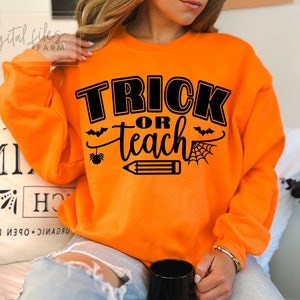 Può includere: Felpa arancione con testo nero che recita "Trick or Teach" con una grafica di pipistrello, ragno, matita e ragnatela.