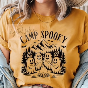 May include: Mustard yellow t-shirt with a black graphic that says "Camp Spooky". The graphic features smiling ghosts around a campfire, with mountains, trees, and houses in the background. A bat is flying above the mountains.