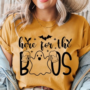Könnte beinhalten: Ein senfgelbes T-Shirt mit einer schwarzen Grafik eines Geistes, der zwei Bierkrüge hält. Der Text "here for the boos" ist auf das T-Shirt gedruckt.