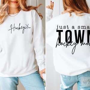 Może przedstawiać: Biała bluza z czarnym napisem z przodu, kt&oacute;ry brzmi "Hockey" i czarnym napisem z tyłu, kt&oacute;ry brzmi "Just a small town hockey mom" z małym sercem.