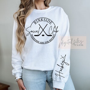 May include: White sweatshirt with a black circle graphic that says "rinkside social club" with hockey sticks crossed inside the circle. The text below the circle says "weekends are for hockey". The left sleeve has the word "hockey" written in cursive.