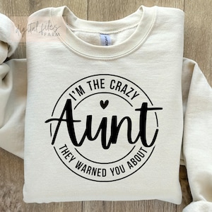 Könnte beinhalten: Ein cremefarbener Pullover mit einem schwarzen Kreis-Motiv, auf dem "I'm the Crazy Aunt They Warned You About" mit einem kleinen schwarzen Herz in der Mitte steht.