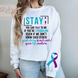 K&ouml;nnte beinhalten: Wei&szlig;er Pullover mit einem t&uuml;rkisfarbenen und violetten Band am &Auml;rmel. Der Text auf dem Pullover lautet "STAY; YOU CAN TALK TO ME IF YOU'RE STRUGGLING (EVEN IF WE DON'T KNOW EACH OTHER) your story is not over your life matters".