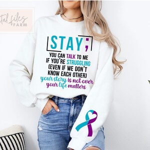K&ouml;nnte beinhalten: Wei&szlig;er Pullover mit dem Text "STAY; YOU CAN TALK TO ME IF YOU'RE STRUGGLING (EVEN IF WE DON'T KNOW EACH OTHER) your story is not over your life matters". Der Pullover hat ein t&uuml;rkisfarbenes und violettes Band am &Auml;rmel.
