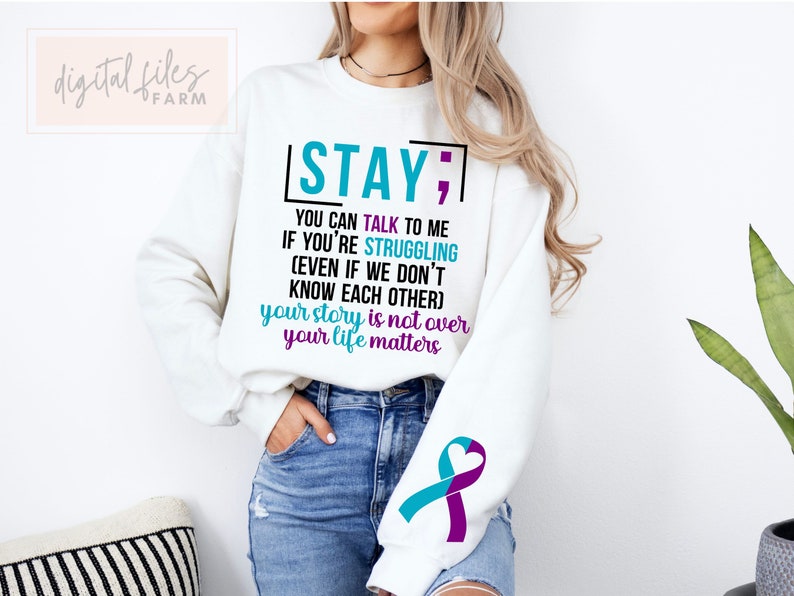 K&ouml;nnte beinhalten: Wei&szlig;er Pullover mit dem Text "STAY; YOU CAN TALK TO ME IF YOU'RE STRUGGLING (EVEN IF WE DON'T KNOW EACH OTHER) your story is not over your life matters". Der Pullover hat ein t&uuml;rkisfarbenes und violettes Band am &Auml;rmel.