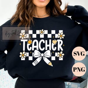 May include: A black long-sleeve sweatshirt featuring a white checkered pattern background with the word TEACHER in white letters. The design includes yellow daisy flowers, a small yellow star, a heart, and a white bow tied around a pencil.