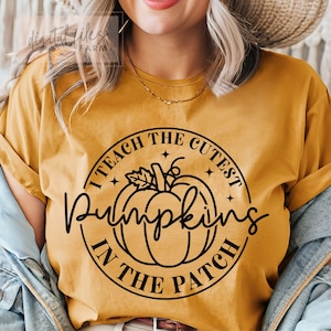 May include: A mustard yellow t-shirt with a black circle graphic that says "I teach the cutest pumpkins in the patch". The graphic features a stylized pumpkin with a stem and leaves.