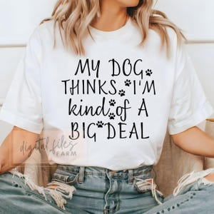 Op de afbeelding: Wit T-shirt met zwarte tekst die "My Dog Thinks I'm Kind of a Big Deal" luidt met poot afdrukken tussen de woorden.