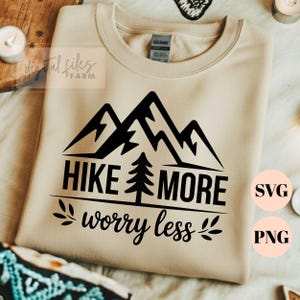 Peut inclure: Un sweat-shirt beige avec un motif de montagne noir et le texte "HIKE MORE worry less". Le design comprend un arbre et des feuilles décoratives. L'étiquette de la marque Gildan est visible. L'image comprend également des icônes de type de fichier SVG et PNG.