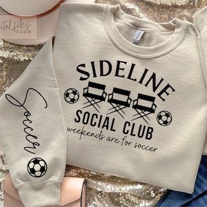 Puede incluir: Una sudadera beige con el texto "Sideline Social Club" y "Weekends are for soccer" en negro. El diseño también incluye tres sillas de director y un balón de fútbol.