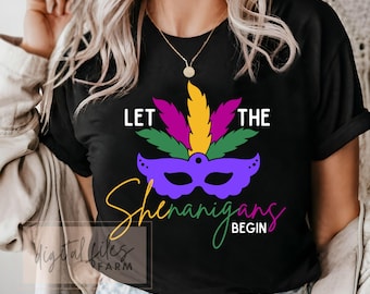 May include: Black t-shirt with a colorful Mardi Gras mask design. The shirt features the text "LET THE SHENANIGANS BEGIN" in white, yellow, and green script. A purple mask with green, yellow, and pink feathers is in the center.