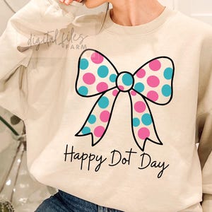 Könnte beinhalten: Beiges Sweatshirt mit einem großen Schleifendesign. Der Bogen hat rosa und türkisfarbene Tupfen, die schwarz umrandet sind. Unter der Schleife steht der Text "Happy Dot Day" in schwarzer Schrift. Die Worte "digital files farm" sind in der oberen linken Ecke.