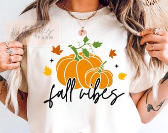 Może przedstawiać: Kremowa koszulka z grafiką dwóch pomarańczowych dyni z zielonymi pędami i liśćmi oraz napisem "fall vibes" czarną czcionką. Wokół dyni rozrzucone są pomarańczowe i żółte liście.