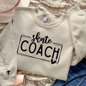 Op de afbeelding: Een beige sweatshirt met zwarte tekst die "skate COACH!" en een klein hartje zegt. De linkermouw heeft de tekst "All Day. Every. Day." en een kleine afbeelding van een schaats.