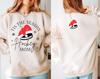 May include: A cream-colored sweatshirt with the text "Tis the Season Hockey Mom" and a red and white hockey puck with a Santa hat on top. The back of the sweatshirt has a single hockey puck with a Santa hat on top.