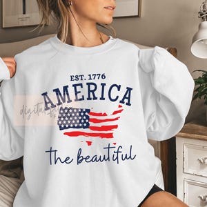 May include: White sweatshirt with a blue and red American flag graphic and the text "EST. 1776 AMERICA the beautiful".