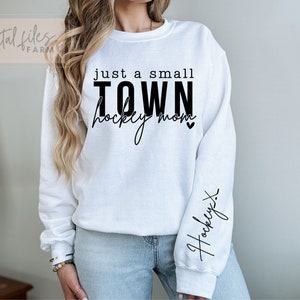 Może przedstawiać: Biała bluza z czarnym napisem "just a small TOWN hockey mom" z małym sercem. Na prawym rękawie napis "hockey" napisany kursywą.