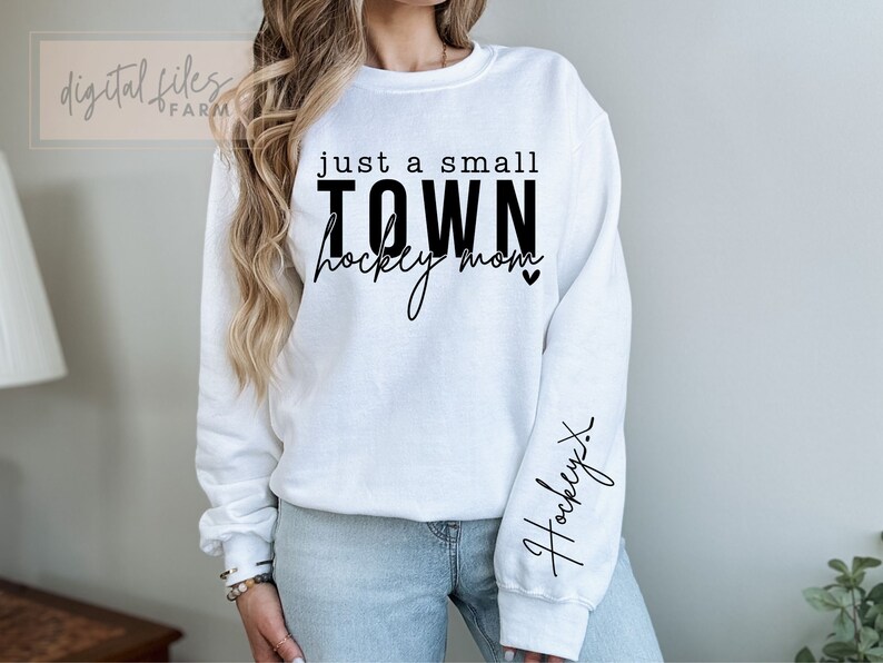 Może przedstawiać: Biała bluza z czarnym napisem "just a small TOWN hockey mom" z małym sercem. Na prawym rękawie napis "hockey" napisany kursywą.