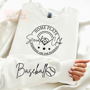 May include: White sweatshirt with a black graphic design that says "Home Plate Social Club Weekends Are For Baseball" with a baseball diamond and three stars. The bottom of the sweatshirt has a black graphic design that says "Baseball" with a baseball.