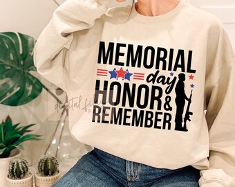 May include: A beige sweatshirt with the words "MEMORIAL day HONOR & REMEMBER" in black, red, and blue. A silhouette of a soldier holding a rifle is on the right side of the text. The sweatshirt is worn with blue jeans.