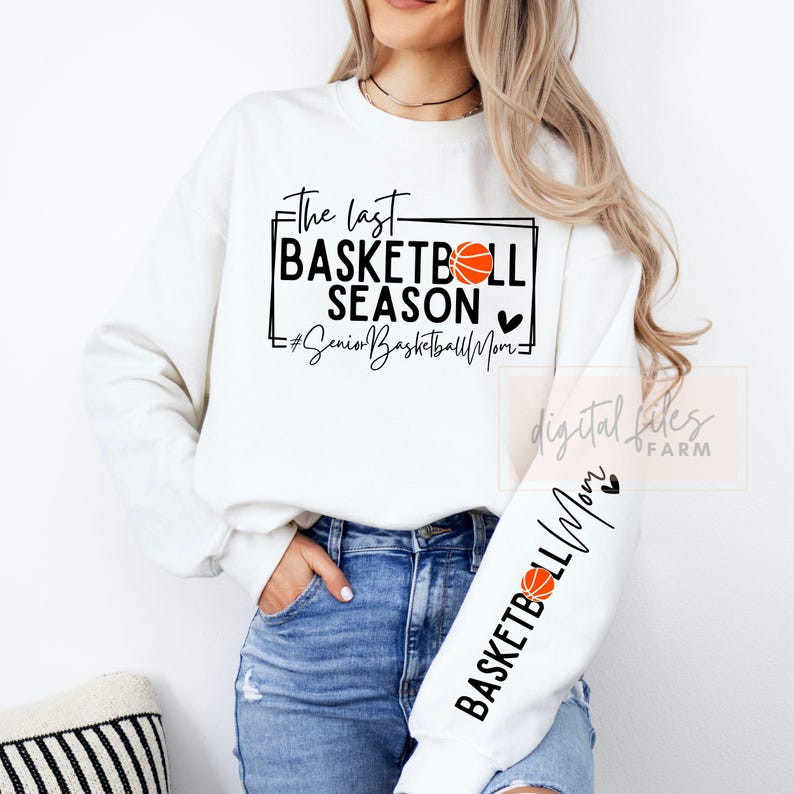 May include: White sweatshirt with black text that reads "The last basketball season #SeniorBasketballMom" with a basketball graphic. The sweatshirt has a black basketball graphic on the sleeve with the text "Basketball Mom" and a heart.