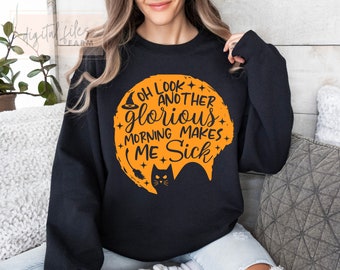 Op de afbeelding: Zwart sweatshirt met een oranje afbeelding van een heksenhoed, een wassende maan en een zwarte kat. De tekst op de afbeelding luidt "Oh look another glorious morning makes me sick."