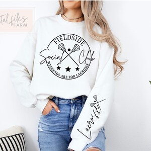 Może przedstawiać: Biała bluza z czarnym kołem graficznym, na kt&oacute;rym widnieje napis "Fieldside Social Club" z krzyżującymi się kijami do lacrosse i tekst "Weekends are for Lacrosse". Lewy rękaw ma napis "Lacrosse" napisany kursywą.