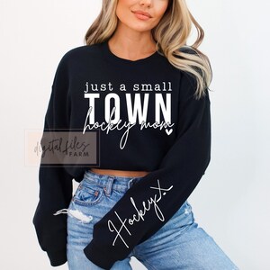 Może przedstawiać: Czarna, kr&oacute;tka bluza z nadrukiem "just a small TOWN hockey mom" w kolorze białym. Bluza ma długi rękaw z napisem "hockey" w kolorze białym na rękawie.
