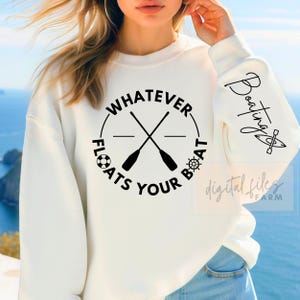 Könnte beinhalten: Weißer Pullover mit einem schwarzen Kreis-Motiv, auf dem "Whatever Floats Your Boat" mit gekreuzten Rudern und einem Schiffsrad steht. Der Text "Boating" ist in Kursivschrift auf dem Ärmel geschrieben.